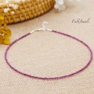 Puede incluir: Un delicado collar de piedras preciosas de color púrpura con un cierre de plata. El collar está hecho de pequeñas cuentas redondas y se muestra sobre un fondo de tela marrón.