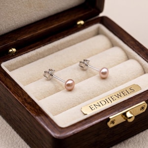 May include: A pair of pink pearl stud earrings displayed in a wooden jewelry box with a cream-colored interior. The earrings are set on silver posts. The box has a gold-colored clasp and the text "ENDIJEWELS" on a gold-colored plate.