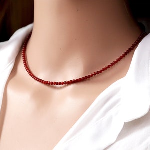 May include: A delicate red beaded necklace. The necklace is composed of small, round beads, creating a simple yet elegant design. The necklace is worn against a white shirt.