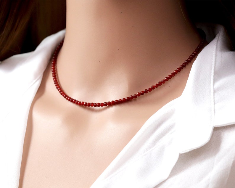 May include: A delicate red beaded necklace. The necklace is composed of small, round beads, creating a simple yet elegant design. The necklace is worn against a white shirt.