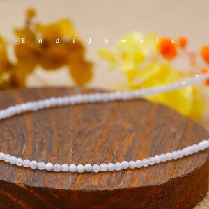 May include: A delicate, light blue beaded necklace. The necklace is displayed on a wooden surface with the text "Endi Jewels" in the background. The background also features blurred yellow and orange flowers.