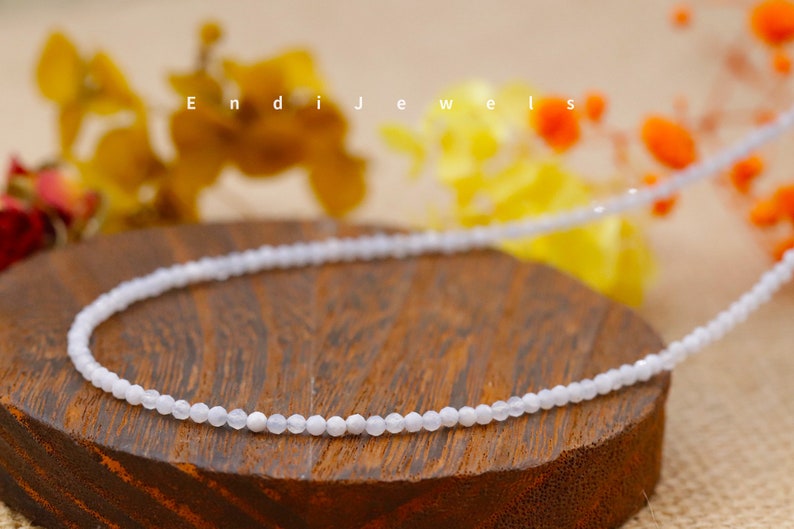 May include: A delicate, light blue beaded necklace. The necklace is displayed on a wooden surface with the text "Endi Jewels" in the background. The background also features blurred yellow and orange flowers.