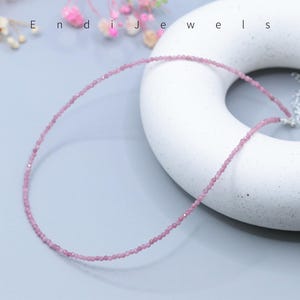 May include: A delicate pink gemstone necklace. The necklace is composed of small, round, light pink beads strung together. The necklace is displayed on a white, donut-shaped object. The word "Endi Jewels" is visible in the background.