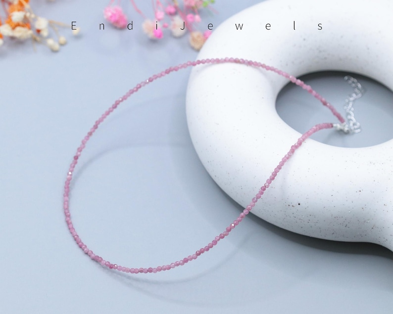 May include: A delicate pink gemstone necklace. The necklace is composed of small, round, light pink beads strung together. The necklace is displayed on a white, donut-shaped object. The word "Endi Jewels" is visible in the background.