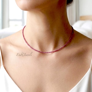 Puede incluir: Un delicado collar de cuentas de color rubí. El collar se lleva alrededor del cuello, con una parte superior blanca en el fondo. La marca "Endi Jewels" es visible en la parte inferior izquierda de la imagen.