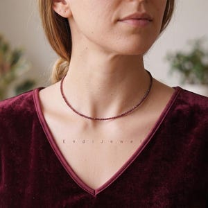 May include: A fine, burgundy beaded necklace. The necklace is worn by a person wearing a burgundy velvet top. The text "Endi Jewel" is visible on the top. The necklace is a choker style.