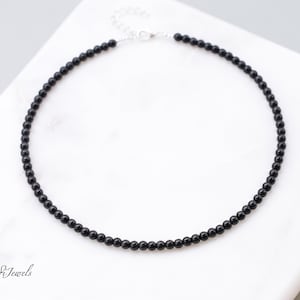 Puede incluir: Un collar de cuentas negras con cierre plateado. El collar est&aacute; compuesto por peque&ntilde;as cuentas negras redondas ensartadas. El collar se exhibe sobre una superficie de m&aacute;rmol blanco.