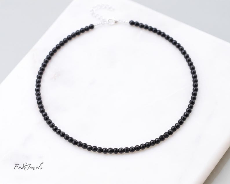 Puede incluir: Un collar de cuentas negras con cierre plateado. El collar est&aacute; compuesto por peque&ntilde;as cuentas negras redondas ensartadas. El collar se exhibe sobre una superficie de m&aacute;rmol blanco.