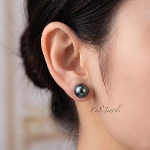May include: A close-up of a dark gray pearl stud earring. The pearl has a lustrous sheen and is set against the ear of a person. The text "Endi Jewels" is visible in the image.