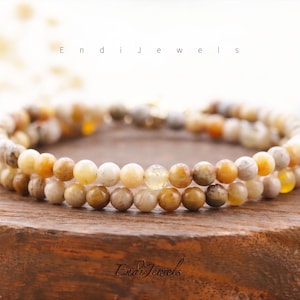 May include: Two beaded bracelets with a mix of beige, brown, and yellow tones. The bracelets are made of small, round beads and are displayed on a wooden surface. The text "Endi Jewels" is visible.
