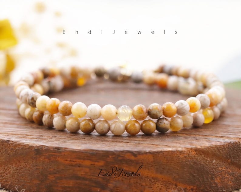 May include: Two beaded bracelets with a mix of beige, brown, and yellow tones. The bracelets are made of small, round beads and are displayed on a wooden surface. The text "Endi Jewels" is visible.