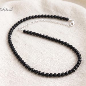 Puede incluir: Un collar de cuentas negras con una cadena y cierre plateados. El collar est&aacute; compuesto por peque&ntilde;as cuentas negras redondas. La joya se exhibe sobre una superficie blanca texturizada. La marca "Endi Jewels" es visible en la esquina superior izquierda.
