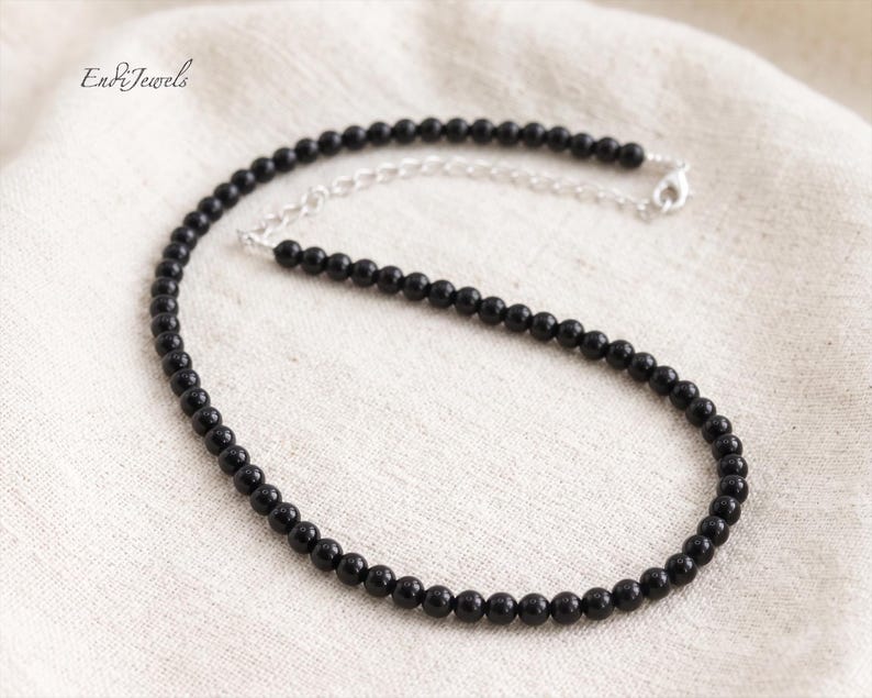 Puede incluir: Un collar de cuentas negras con una cadena y cierre plateados. El collar est&aacute; compuesto por peque&ntilde;as cuentas negras redondas. La joya se exhibe sobre una superficie blanca texturizada. La marca "Endi Jewels" es visible en la esquina superior izquierda.