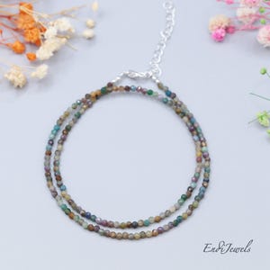 May include: A delicate necklace featuring two strands of small, multi-colored beads. The beads display hues of green, brown, and blue. The necklace has a silver-toned clasp and chain. The brand name "Endi Jewels" is visible in the bottom right corner.