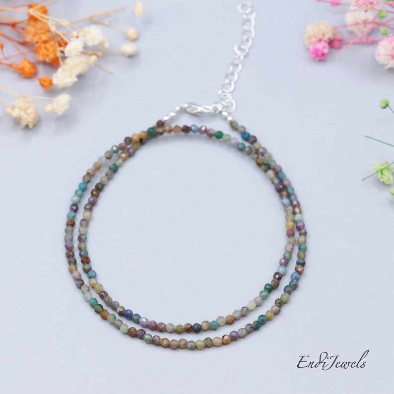 May include: A delicate necklace featuring two strands of small, multi-colored beads. The beads display hues of green, brown, and blue. The necklace has a silver-toned clasp and chain. The brand name "Endi Jewels" is visible in the bottom right corner.