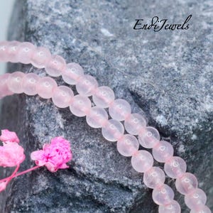 May include: A strand of smooth, translucent pale pink beads arranged in a double loop. The beads are uniform in size and spherical. The text EndiJewels is visible in black script in the upper right corner, alongside small pink flowers.