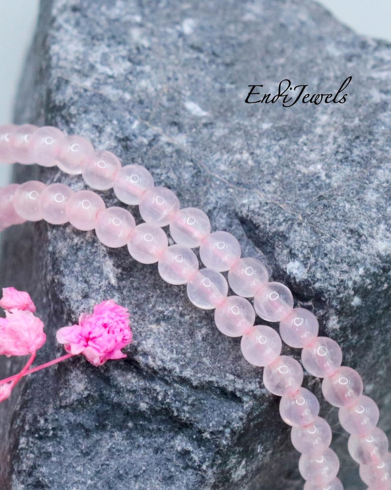 May include: A strand of smooth, translucent pale pink beads arranged in a double loop. The beads are uniform in size and spherical. The text EndiJewels is visible in black script in the upper right corner, alongside small pink flowers.
