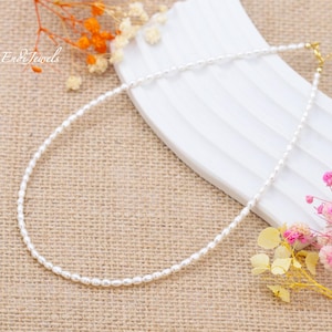 May include: A white pearl necklace with a gold clasp. The necklace is on a beige fabric background with dried flowers.