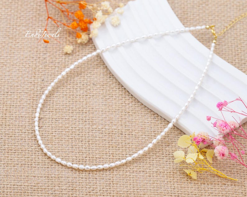 May include: A white pearl necklace with a gold clasp. The necklace is on a beige fabric background with dried flowers.