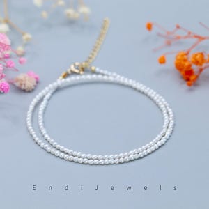 May include: Delicate pearl necklace with a gold clasp, displayed on a light gray surface. The necklace is composed of multiple strands of small, round, white pearls. The word "Endi Jewels" is printed at the bottom of the image. Pink and orange flowers are in the background.