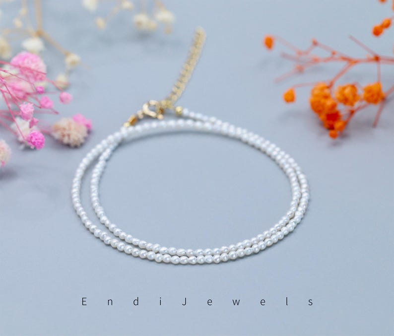 May include: Delicate pearl necklace with a gold clasp, displayed on a light gray surface. The necklace is composed of multiple strands of small, round, white pearls. The word "Endi Jewels" is printed at the bottom of the image. Pink and orange flowers are in the background.