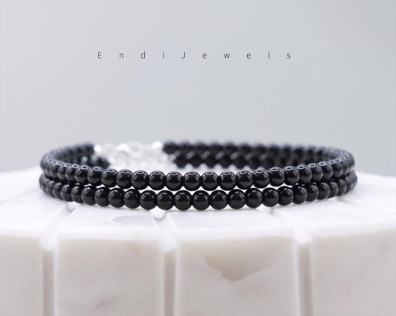 Puede incluir: Una pulsera de dos hilos con peque&ntilde;as cuentas negras brillantes. La pulsera est&aacute; conectada con un cierre plateado. El nombre de la marca "Endi Jewels" se muestra en una fuente minimalista encima de la pulsera.