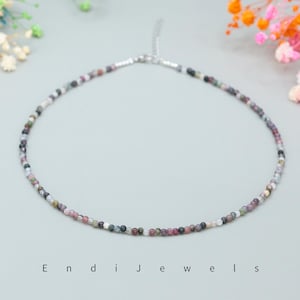 May include: A delicate necklace made of small, multi-colored beads. The beads are a mix of pink, purple, gray, and black. The necklace is on a white background.