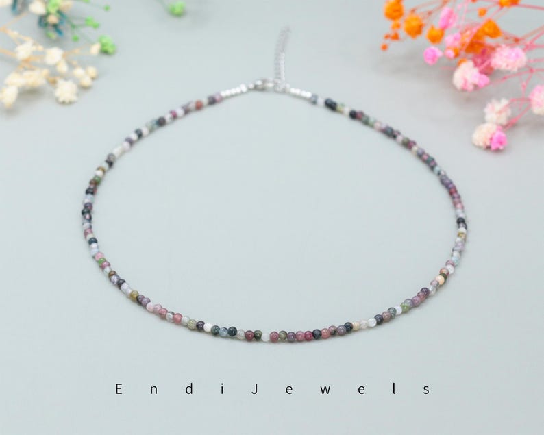 May include: A delicate necklace made of small, multi-colored beads. The beads are a mix of pink, purple, gray, and black. The necklace is on a white background.
