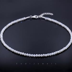 May include: A delicate white beaded necklace with a silver clasp. The necklace is composed of small, round beads and is displayed on a dark reflective surface. The text "E n d i J e w e l s" is at the bottom of the image.