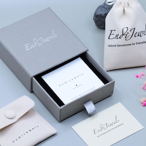 May include: A gray jewelry box with the brand name "Endi Jewels" in silver lettering. The box is open, revealing a white card. A small drawstring bag and a pouch with the brand name are also visible, along with a card that reads "Endi Jewels".