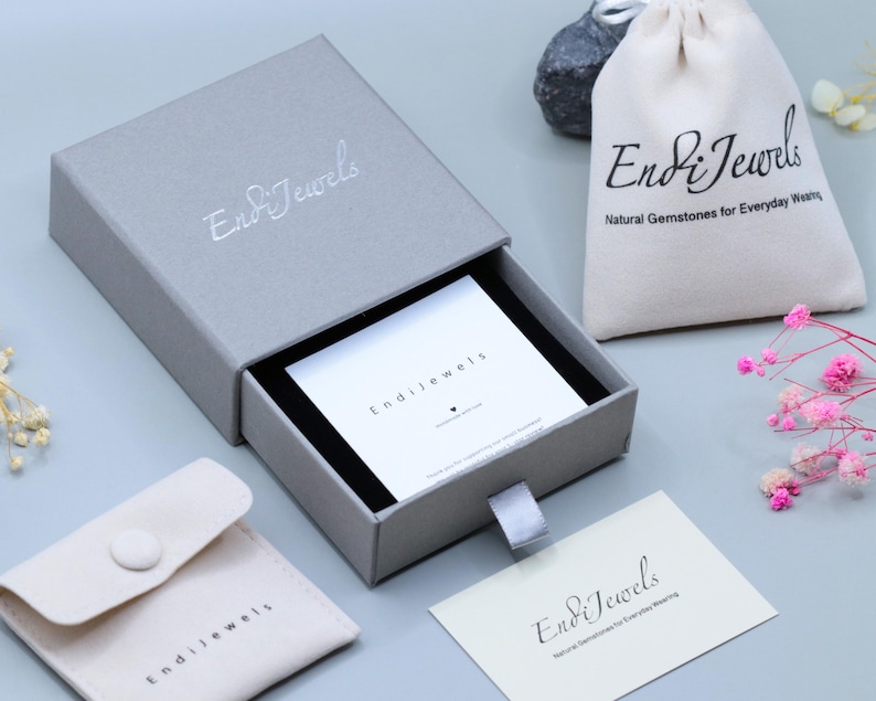 May include: A gray jewelry box with the brand name "Endi Jewels" in silver lettering. The box is open, revealing a white card. A small drawstring bag and a pouch with the brand name are also visible, along with a card that reads "Endi Jewels".