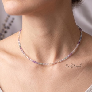 May include: A delicate necklace composed of small, colorful beads in shades of purple, green, and clear. The necklace is worn around the neck. The text "En&Jewels" is visible on the lower right.