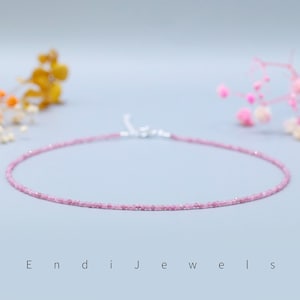 May include: A delicate pink gemstone necklace with a silver clasp. The necklace is composed of small, round, pink gemstones. The background is a soft, light blue with blurred floral accents and the text "Endi Jewels".