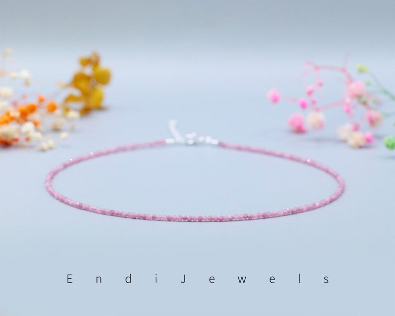 May include: A delicate pink gemstone necklace with a silver clasp. The necklace is composed of small, round, pink gemstones. The background is a soft, light blue with blurred floral accents and the text "Endi Jewels".