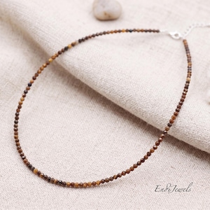 May include: A delicate necklace featuring small, faceted brown beads strung together. The necklace has a silver-colored clasp and is displayed on a neutral-colored fabric. The overall design is simple and elegant, suitable for everyday wear.