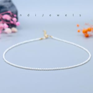 May include: Delicate white beaded necklace with a gold clasp, displayed on a light blue surface. The necklace is a simple, elegant design. The word "Endi Jewels" is written in white letters.