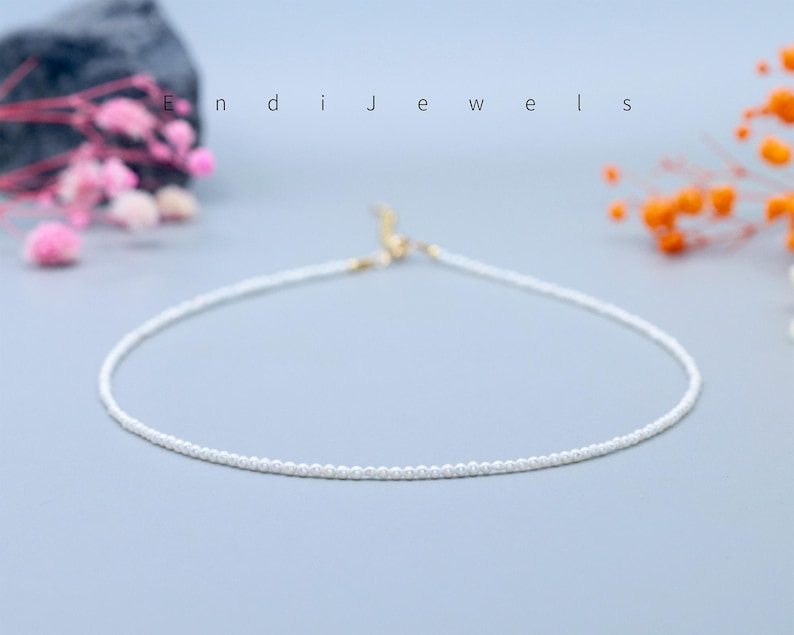 May include: Delicate white beaded necklace with a gold clasp, displayed on a light blue surface. The necklace is a simple, elegant design. The word "Endi Jewels" is written in white letters.
