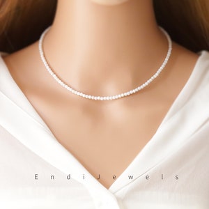 Puede incluir: Un collar de gargantilla simple de perlas blancas sobre un fondo blanco. El collar está hecho de pequeñas perlas redondas y mide aproximadamente 40 cm de largo.