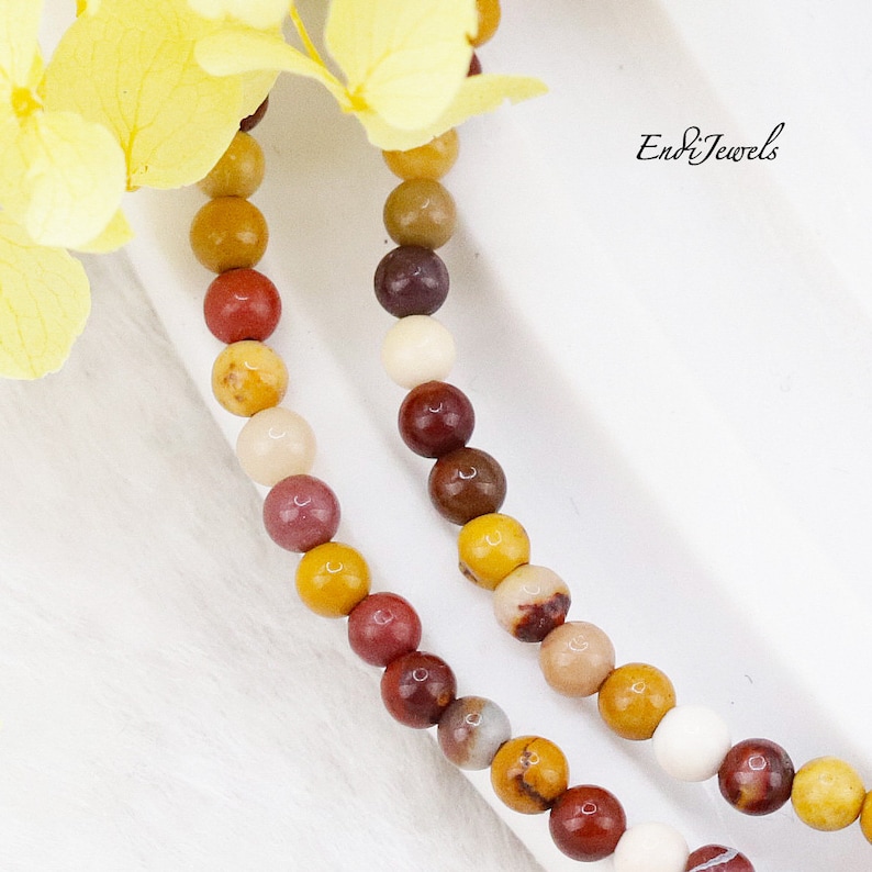 May include: A close-up of a multi-colored beaded necklace with red, brown, yellow, and white beads. The necklace is on a white surface. Endi Jewels is written in the top right corner of the image.