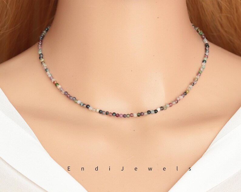 May include: A delicate necklace made of small, multi-colored beads. The beads are a mix of pink, green, and black, and they are strung on a silver chain.