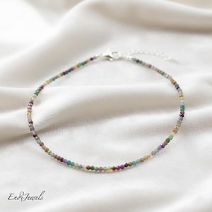 May include: A delicate beaded anklet featuring small, colorful gemstones in shades of purple, green, yellow, and brown. The anklet is strung on a silver chain and rests on a white, silky fabric. The brand name "Endi Jewels" is in the bottom left corner.
