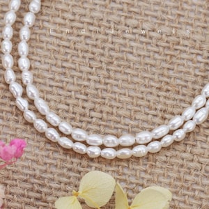 May include: A double strand of white pearl beads on a burlap background. The pearls are oval shaped and have a smooth, glossy finish.