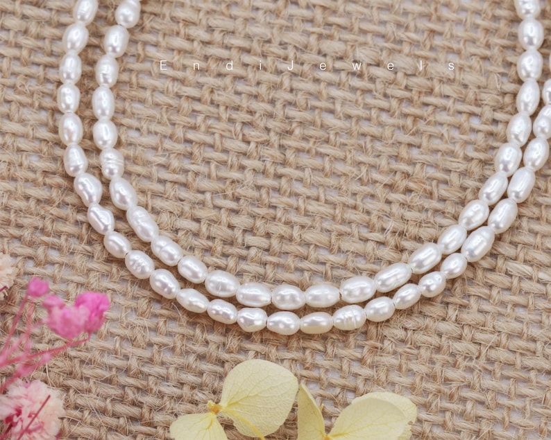 May include: A double strand of white pearl beads on a burlap background. The pearls are oval shaped and have a smooth, glossy finish.