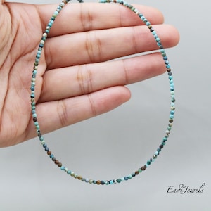 Op de afbeelding: Een delicate turquoise en bruine edelsteen ketting met kleine, ronde kralen. De ketting ligt op een hand, waardoor de lengte en grootte van de kralen zichtbaar zijn.