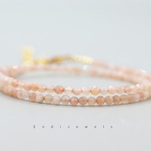 K&ouml;nnte beinhalten: Ein zartes Armband mit zwei Reihen aus kleinen, hellrosa Sunstone-Perlen. Das Armband hat einen goldenen Verschluss.