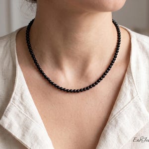 Puede incluir: Un collar de cuentas negras sobre el cuello de una persona. El collar est&aacute; compuesto por peque&ntilde;as cuentas negras redondas. La persona lleva una camisa de lino de color claro. El nombre de la tienda Etsy "Endi Jewels" es visible en la esquina inferior derecha.
