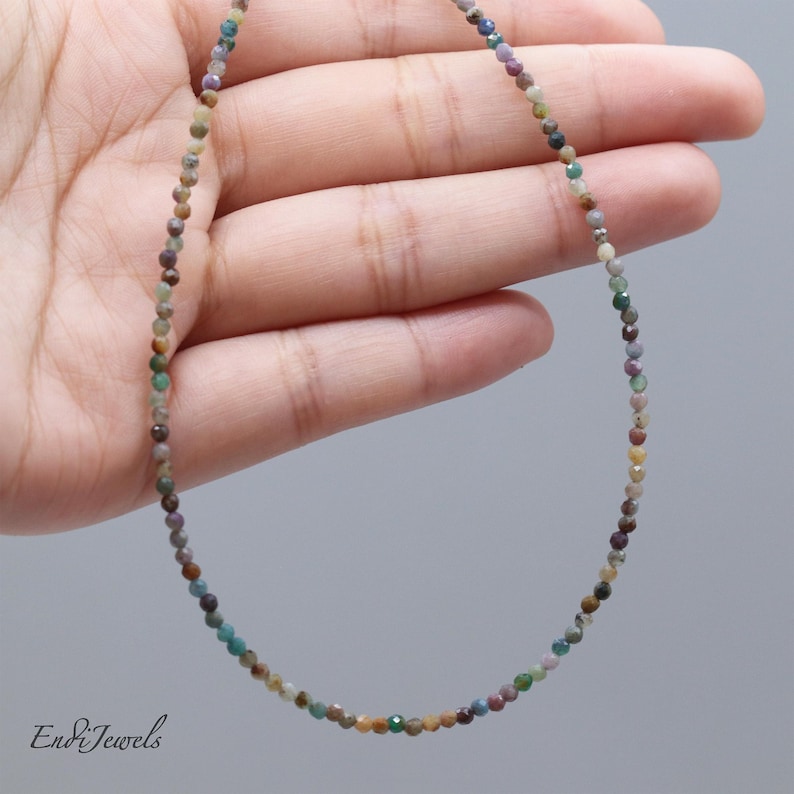 May include: A delicate necklace composed of small, multi-colored beads. The beads are in shades of green, brown, purple, and yellow. The necklace is held in a hand against a gray background. The text "Endi Jewels" is in the bottom left corner.