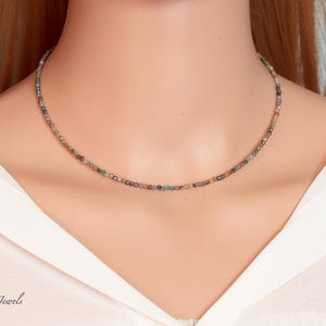 May include: A delicate necklace composed of small, multi-colored beads. The beads display hues of green, brown, and red, strung together to form a choker-style necklace. The necklace is worn against a white shirt.