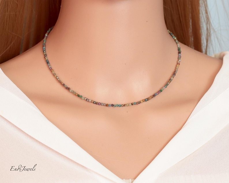 May include: A delicate necklace composed of small, multi-colored beads. The beads display hues of green, brown, and red, strung together to form a choker-style necklace. The necklace is worn against a white shirt.
