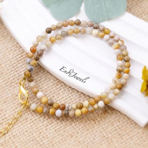 May include: A delicate necklace featuring small, round beads in shades of brown, tan, and cream. The necklace has a gold-colored clasp and chain. The text "End & Jewels" is visible on the white surface.